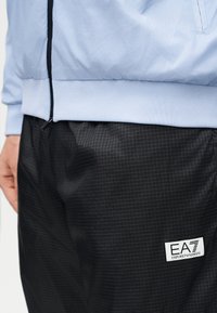 Κοντινό πλάνο ατόμου που φοράει μαύρο καρό παντελόνι EA7 Emporio Armani και ανοιχτό μπλε μπουφάν με φερμουάρ και ελαστική μέση.