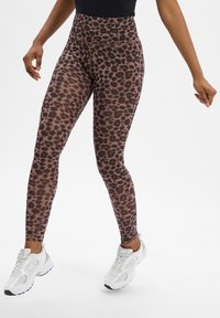 Legginsy z wysokim stanem w brązowo-czarne w leopardzie, wykonane z elastycznego materiału. Połączone z białymi butami sportowymi. Gładka tekstura.
