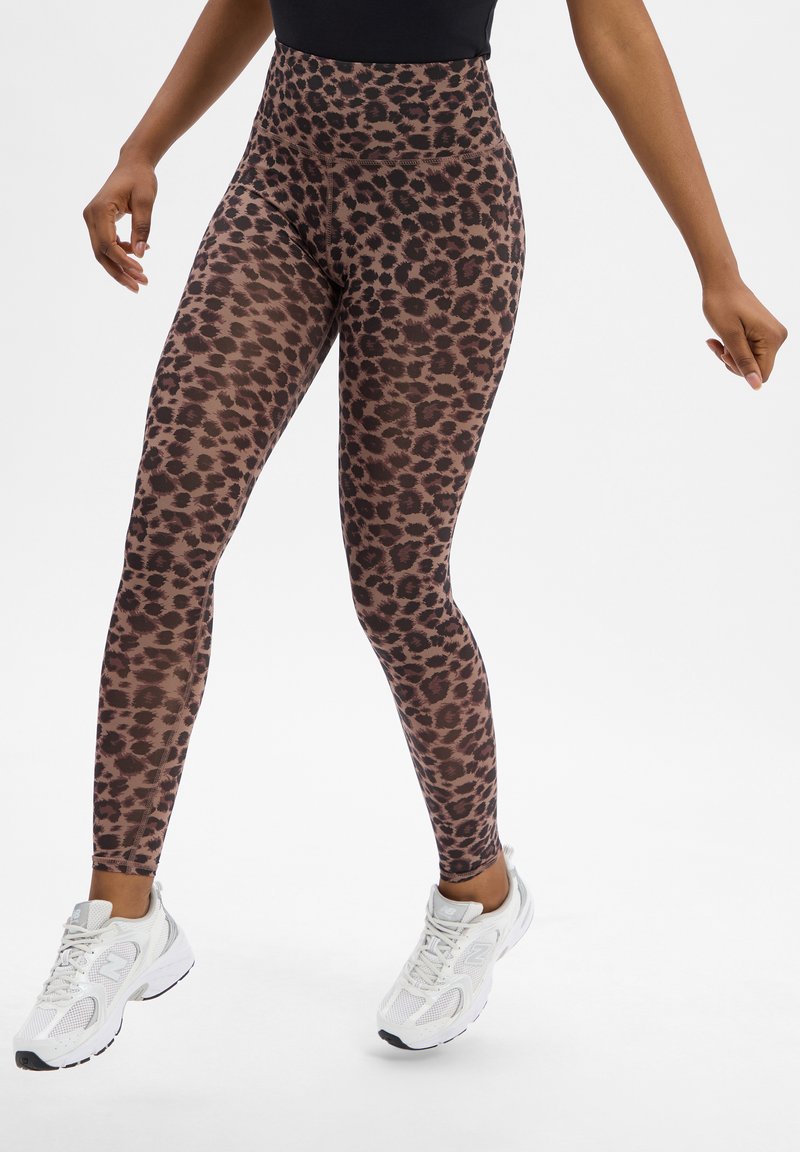 Legginsy z wysokim stanem w brązowo-czarne w leopardzie, wykonane z elastycznego materiału. Połączone z białymi butami sportowymi. Gładka tekstura.