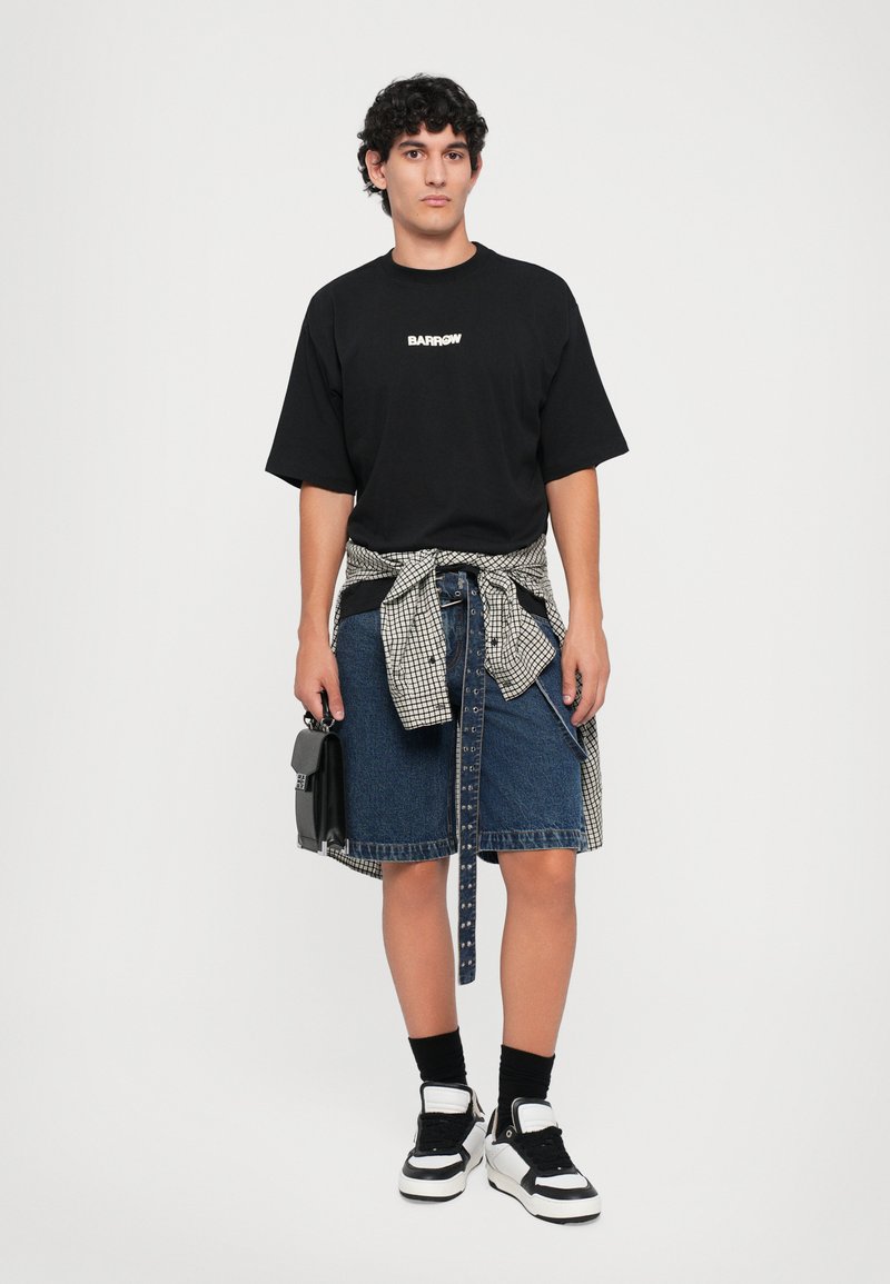 Giovane uomo che indossa una maglietta nera oversize con logo, una gonna di jeans fino al ginocchio, una camicia a quadri annodata in vita, sneakers e tiene una borsa nera.