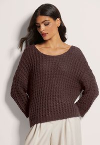 Brauner Strickpullover mit strukturiertem Muster, weitem Ausschnitt und langen Ärmeln, kombiniert mit hellen Hosen. Weicher, gemütlicher Stoff.
