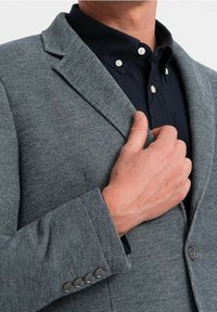 Szary marynarka wykonana z fakturowanej tkaniny, z klapami, pojedynczym zapięciem na guzik i mankietami na cztery guziki, noszona na ciemnej koszuli z kołnierzykiem.