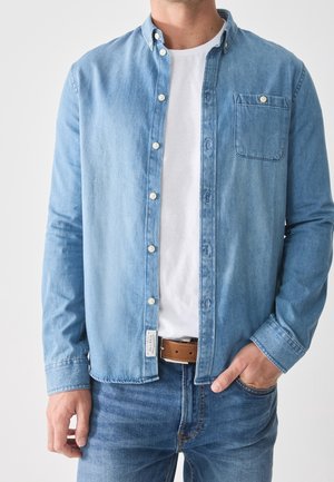 Mężczyzna w jasnoniebieskiej dżinsowej koszuli zapinanej na guziki, na białej koszulce, w niebieskich dżinsach i brązowym pasku, z ręką w kieszeni.