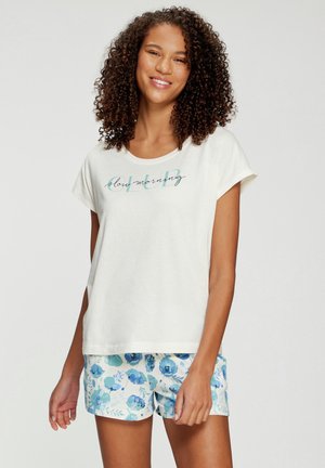 Frau mit lockigem Haar, die ein weißes T-Shirt mit dem Text "slow morning" und blaue Shorts mit Blumenmuster trägt, lächelt vor neutralem Hintergrund.
