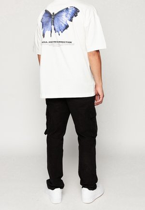 White t-shirt with a large blue butterfly graphic on the back, paired with black cargo pants and white sneakers. Simple design, casual fit.