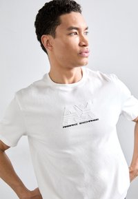 Valge puuvillane T-särk, millel on tõstetud logo "AX" horisontaalsete joontega ja must tekst "ARMANI EXCHANGE". Ümmargune kaelus.