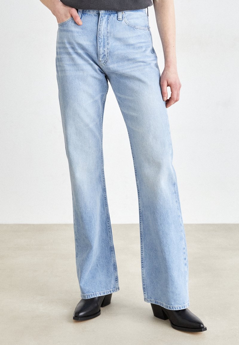 Personne portant un jean large taille haute bleu clair avec des bottines à talons noires, debout avec une main dans la poche sur un fond uni.