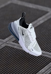 Bela atletska športna obutev z mrežastim zgornjim delom, črnimi poudarki in Air Max oblazinjenjem. Ima teksturiran podplat in detajle z logotipom.