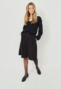 Camisola preta de manga longa combinada com calções pregueados pretos de cintura alta. Mocassins pretos complementam o visual, com meias pretas transparentes.