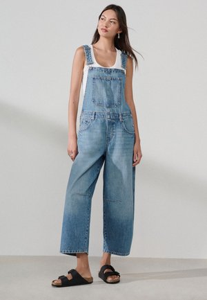 Femme en salopette en denim bleu à jambes larges et débardeur blanc, debout, portant des sandales slides noires sur un fond clair uni.