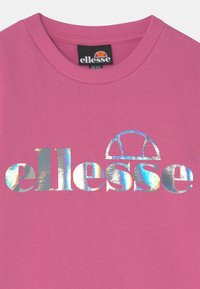 Růžová mikina s žebrovaným límcem a velkým duhově lesklým logem „ellesse“ uprostřed na hrudi. Měkká, texturovaná látka.