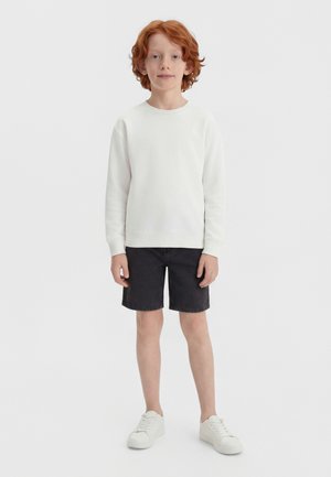 Punapea poiss, kes kannab valget pikkade varrukatega särki, musti lühikesi pükse ja valgeid tossusid, seisab tavalise valge tausta ees.