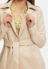 Beige mokkanahkainen takki, jossa vyötäröllä vyö, kauluksessa lovi ja sivutaskut, päällä vihreä kukkakuvioinen paita. Pehmeä tekstuuri, räätälöity malli.