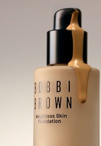 Bobbi Brown Weightless Skin Foundation i en beige flaske med en svart pumpe. Foundation renner fra dysen, noe som viser teksturen.
