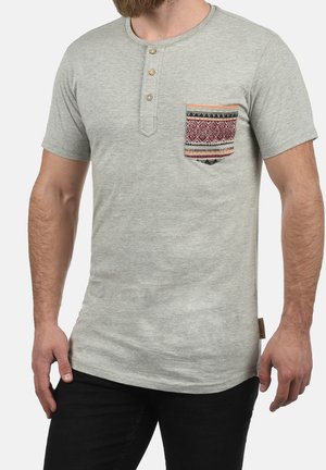 Harmaa lyhythihainen t-paita, jossa on henley-kaulus, neljä nappia ja koristekoti, jossa on punamusta monikuvioinen kangas.