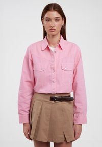 Helles rosa Hemd mit zwei Fronttaschen und langen Ärmeln, kombiniert mit einem khakifarbenen Faltenrock und einem braunen Gürtel. Einfaches und strukturiertes Design.