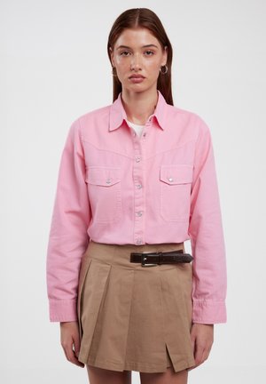 Camicetta button-up di colore rosa chiaro con due tasche frontali e maniche lunghe, abbinata a una gonna plissettata color khaki e una cintura marrone. Design semplice e strutturato.