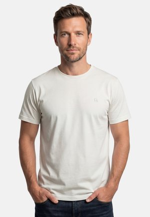 Mann mit kurzem braunem Haar und Bart, trägt ein schlichtes weißes kurzärmliges T-Shirt und dunkle Jeans, Hände in den Taschen, schaut nach vorne.