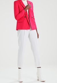 Jasně růžové sako na míru se dvěma knoflíky, doplněné bílými kalhotami s tenkými pruhy a lesklými bílými kotníkovými botami. Hladká látka, moderní střih.