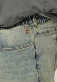 Ljusblå denimjeans med en avslappnad passform, som har en brun logotyp på bakfickan och resårmidja. Lätt urtvättad textur.
