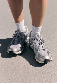 Zapatillas deportivas plateadas que cuentan con una parte superior de malla texturizada, suela de goma y un patrón de tracción negro contrastante. Llevadas con calcetines blancos hasta la mitad de la pierna.