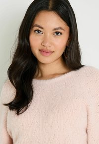 Pull en rose clair, texturé, avec une finition douce et duveteuse et un col bateau, présentant un motif subtil et une coupe décontractée.