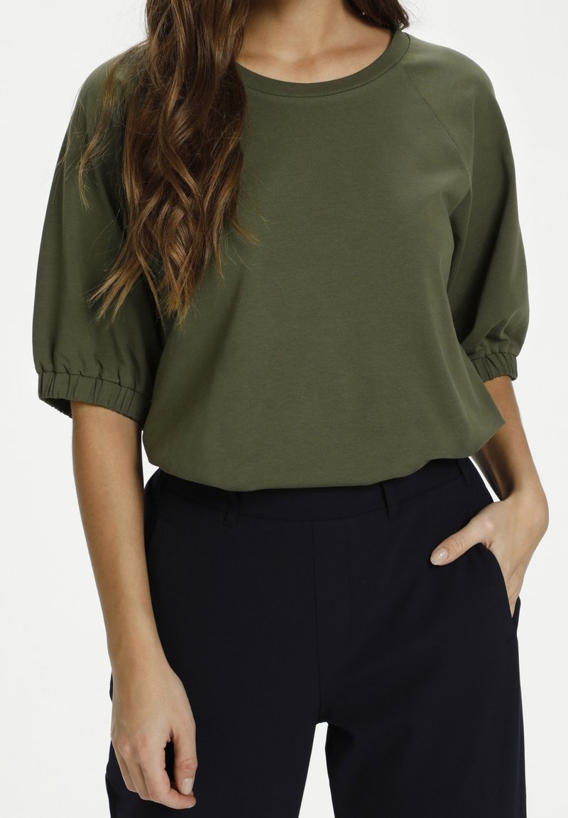 Vrouw in een olijfgroene top met pofmouwen, ingestopt in hoge zwarte broek, met één hand in de zak en lange golvende haren zichtbaar.