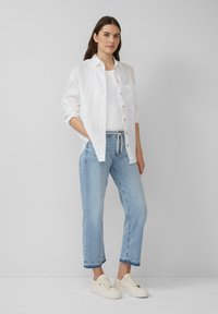 Weißes, knopfbares Leinenhemd über einem weißen Tanktop; hellblaue, lässig geschnittene Jeans mit Kordelzug; crèmefarbene Sneakers.