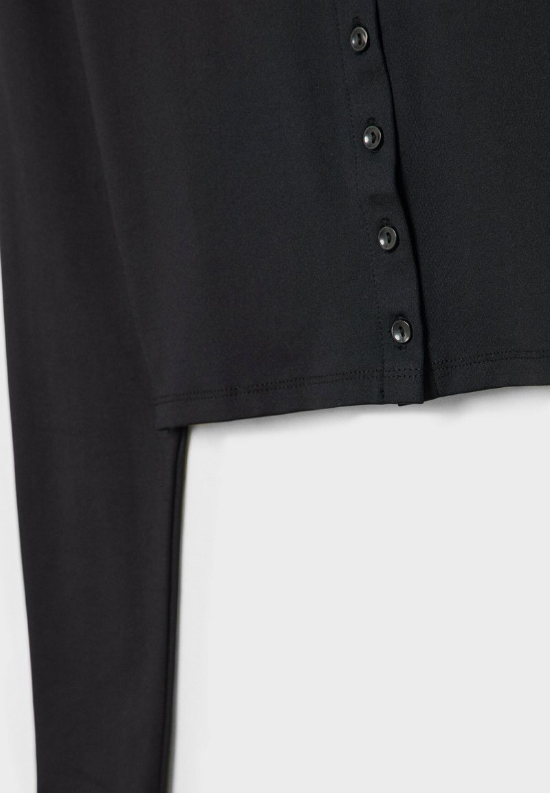 Haut noir à manches longues en tissu doux, avec un ourlet droit et une rangée de boutons noirs sur le devant pour la fermeture.