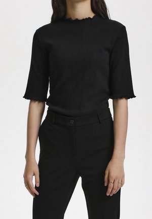 Kobieta ubrana w dopasowaną czarną bluzkę z falistymi brzegami na rękawach i kołnierzu, w połączeniu z czarnymi, wysokowatymi spodniami.