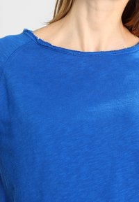 Top blu lungo con maniche lunghe e scollatura ampia, realizzato in un morbido tessuto texturizzato. Presenta bordi sfilacciati all'inguine e sulle spalle.