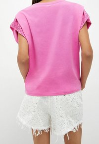 Lyserød kortærmet skjorte med blonder detaljer ved ærmerne, parret med hvide denim shorts prydet med sølvnitter og slidte kanter.