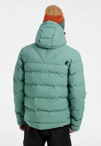 Chaqueta acolchada verde con capucha, que presenta un diseño acolchado y puños elásticos. Detalle del logo en la parte posterior visible. Llevada con un gorro multicolor.