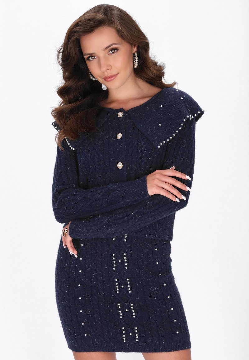 Vestido de punto cableado en color azul marino con un amplio cuello, acentos de botones de perla y perlas decorativas a lo largo del dobladillo y los laterales. Tejido texturizado.