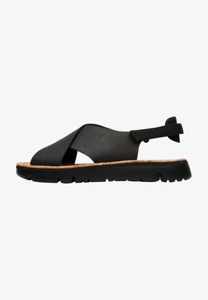 Black sandal with a cross-over design, made of smooth material and a cork sole. Features a textured strap and a chunky rubber base.