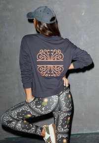 Sudadera corta negra con diseño de remolinos naranjas en la espalda, combinada con leggings negros que presentan un patrón de paisley y sol. Gorra de béisbol gris.
