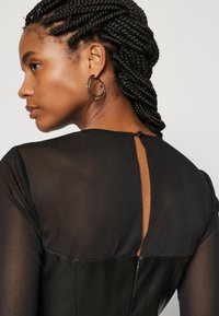 Rochie neagră cu mâneci lungi semi-transparente, corset din piele și o decupare în formă de cheie la spate. Prezentă un închidere cu buton.