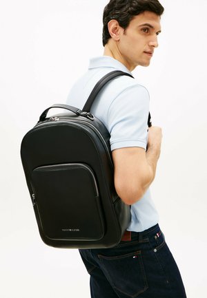 Hombre con camisa tipo polo azul claro y jeans oscuros lleva una mochila de cuero negro con bolsillo frontal con cremallera sobre un hombro.