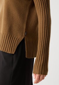 Bruine geribbelde gebreide trui met mouw en zoom, voorzien van een zijsplit, gedragen met zwarte broek op de arm en hand van een persoon.