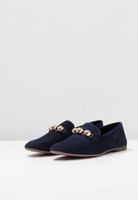Mocasines de ante azul marino con punta afilada y herrajes decorativos en tono dorado en la parte frontal. Suela plana y un acabado suave y mate.