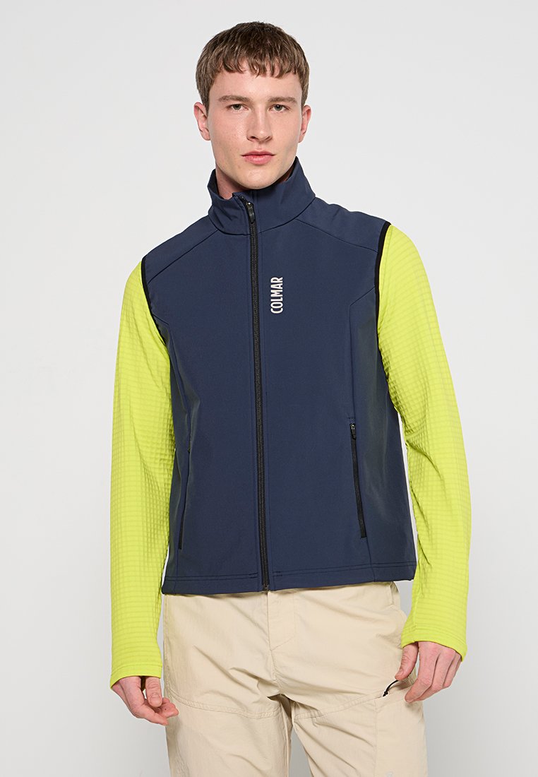 Giovane uomo che indossa un gilet blu navy con zip sopra una maglietta a maniche lunghe verde lime e pantaloni beige, in piedi contro uno sfondo semplice.