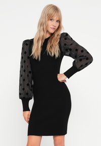 Abito nero a maglia con maniche lunghe trasparenti caratterizzate da pois neri. Silhouette aderente con polsini rigati e scollatura alta.