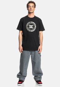 Černé bavlněné tričko s kulatým bílým grafickým logem uprostřed. Nosit s volně střiženými šedými denimovými kalhotami a teniskami s červenými akcenty.