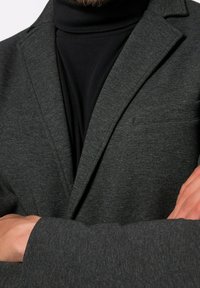 Muž v tmavě šedém saku přes černý rolák, se zkříženýma rukama, s částečně viditelným vousem v horní části rámu.