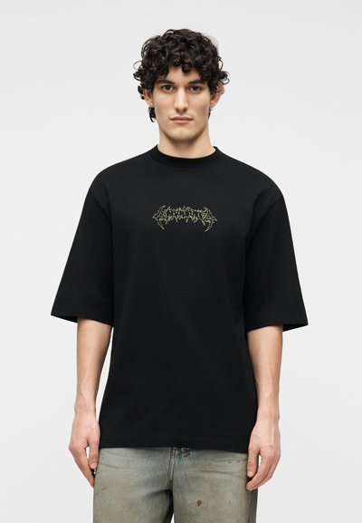 Άνδρας με σγουρά μαλλιά, φορώντας μαύρο υπερμεγέθη T-shirt με περίπλοκη γραφική επιγραφή και ξεθωριασμένο μπλε τζιν, μπροστά σε απλό φόντο.