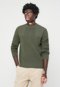 Olive green knit sweater with ribbed texture, crew neckline, and distinct panel detailing. Pairing with beige trousers.