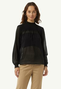Schwarze transparente Bluse mit hohem gestuften Kragen, strukturierten Punkten und langen Ballonärmeln, kombiniert mit beigen Hosen.