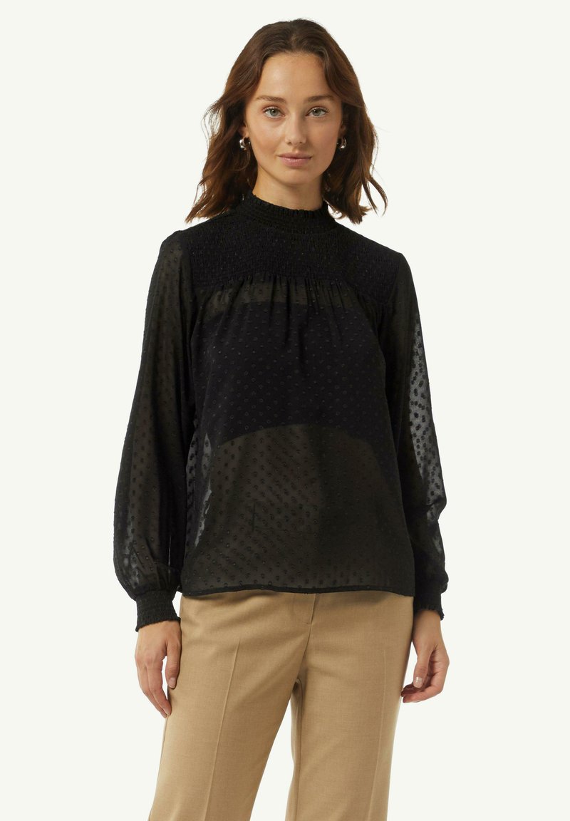 Schwarze transparente Bluse mit hohem gestuften Kragen, strukturierten Punkten und langen Ballonärmeln, kombiniert mit beigen Hosen.