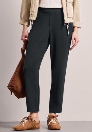 Personne portant un pantalon noir slim, une veste beige, une chemise blanche, des baskets marron, et tenant un sac en daim marron à côté d'elle.