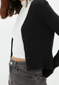 Czarny kardigan prążkowany o dopasowanym kroju, z sześcioma guzikami z przodu. Noszony na białej prążkowanej bluzce z wysokim dekoltem.